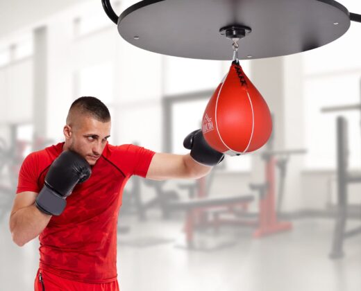 a man in a red t-shirt and red shorts, wearing black boxing gloves, punching a red speed ball in a gym