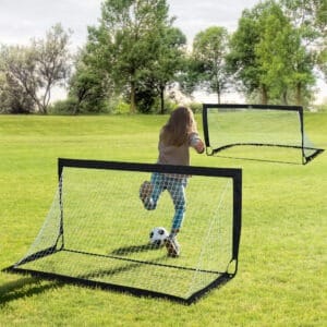 a young girl in a beige top and blue trousers, kicking a ball at an empty goal