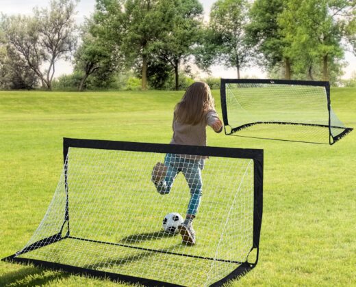 a young girl in a beige top and blue trousers, kicking a ball at an empty goal