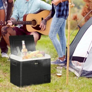 man sitting next to a tent in a field playing a guitar, in front of him is a portable refrigerator filled with fruit and drinks