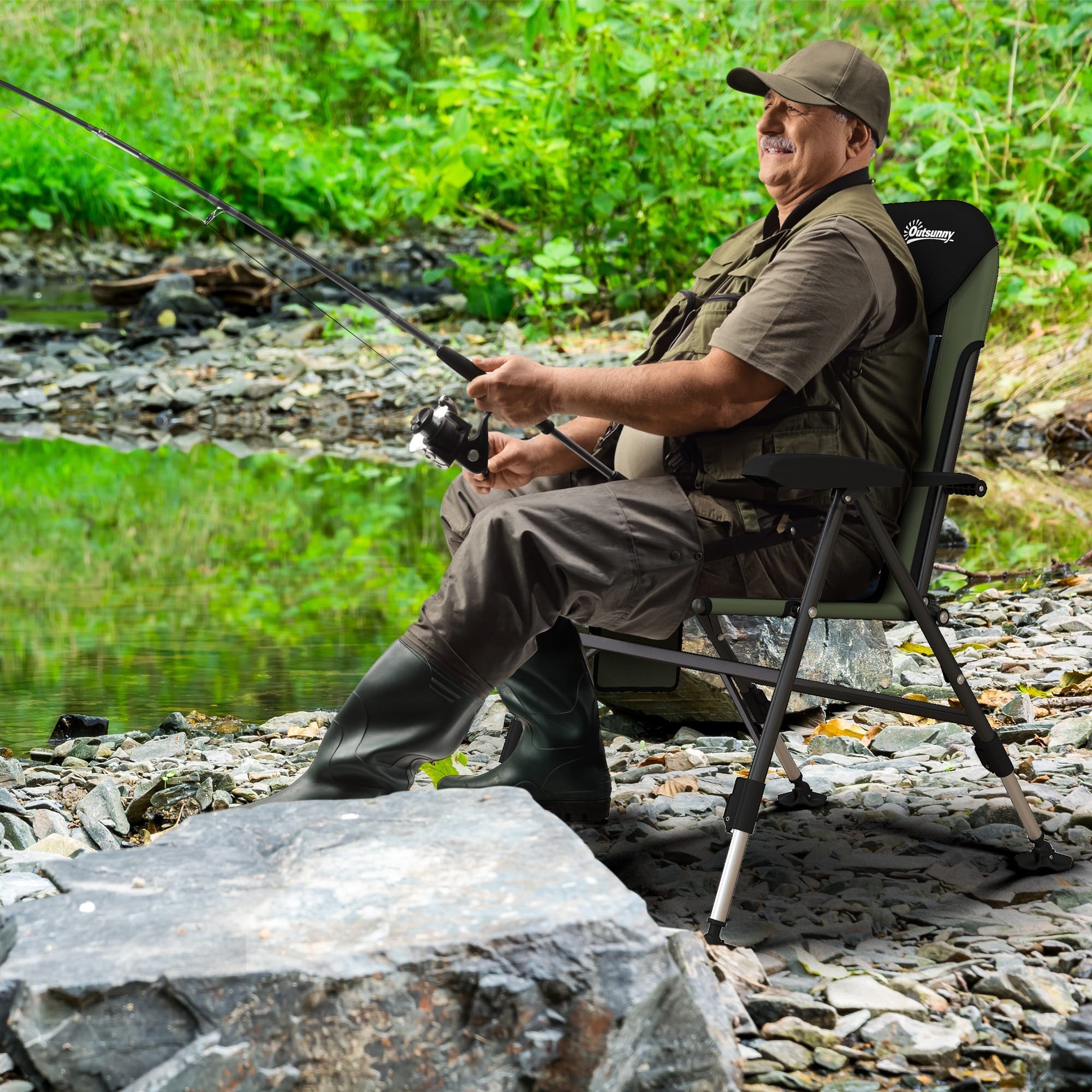 A man in green clothing, a cap and thigh boots, sitting in a chair fishing.