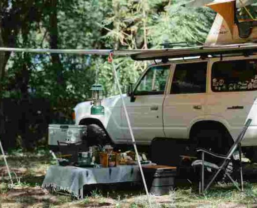 a white 4x4 vehicle, with a tent on the roof and an awning out, covering a camping table and chair, parked in a field surrounded by trees
