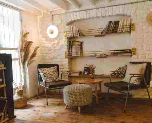 a homely living room with cream painted brick walls, shelving with magazines attached to the wall, two chairs, a coffee table and a footstool on a natural wood floor