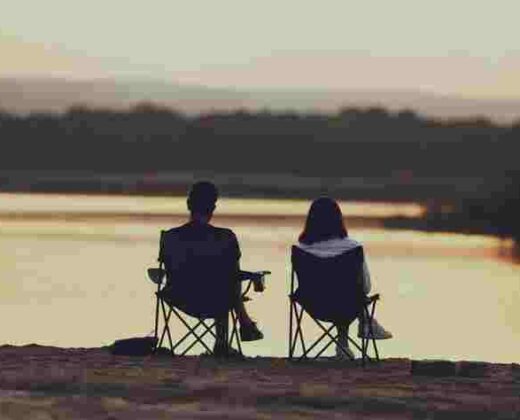 a man and a woman sitting in camping chairs, looking out over a lake at sunset