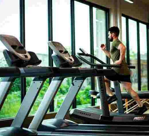 a row of treadmills, in a gym with large glass windows and a man in a green top and black shorts running on a treadmill
