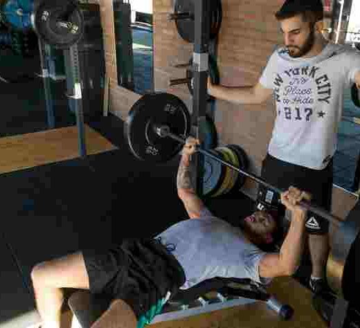 in a gym, a man laying on a weight bench lifting weights with another man overseeing him for safety